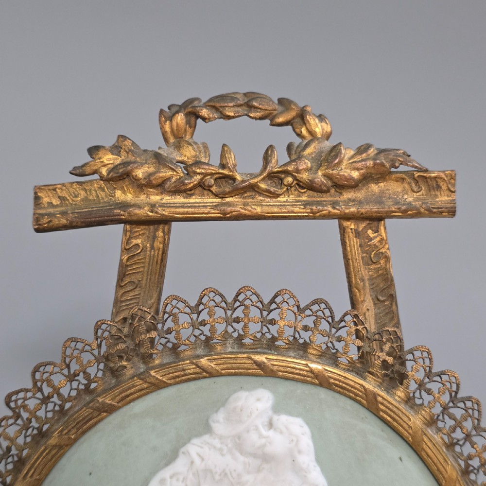 A pale green and white bisque porcelain oval plaque depicting two figures in high relief, supported on an ornate gilt metal easel. View IX