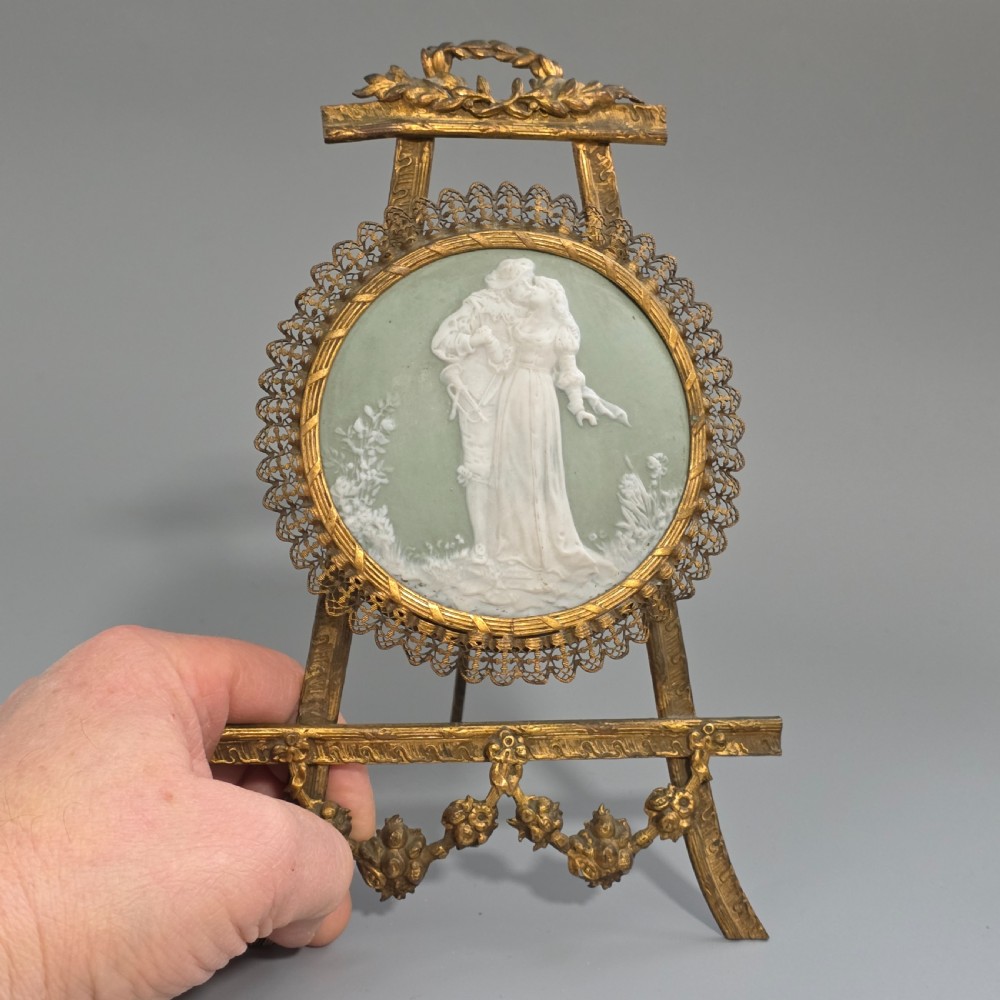 A pale green and white bisque porcelain oval plaque depicting two figures in high relief, supported on an ornate gilt metal easel. View X