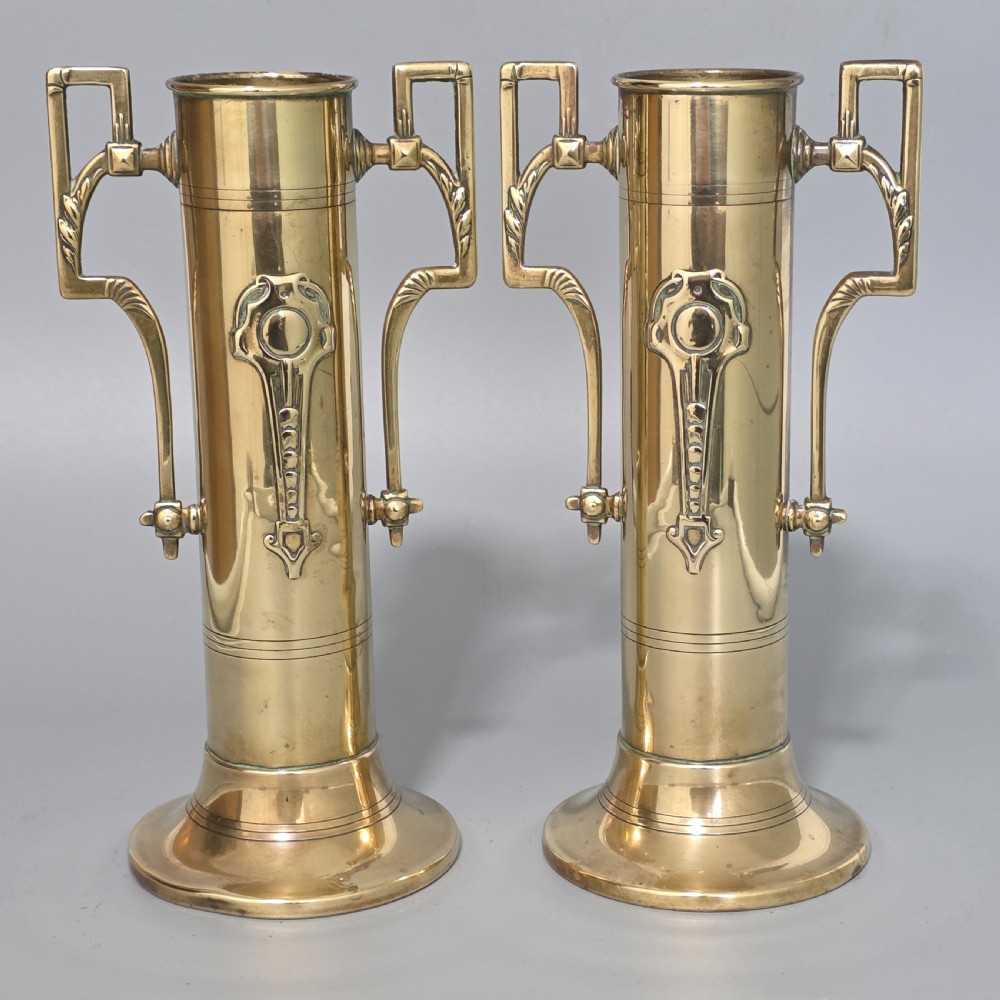 A pair of antique brass cylindrical vases featuring angular twin handles and raised stylised relief details on flared circular bases.