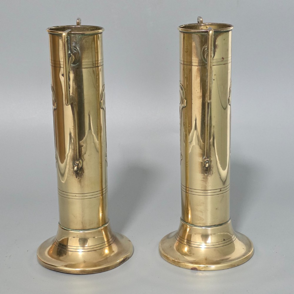 A pair of antique brass cylindrical vases featuring angular twin handles and raised stylised relief details on flared circular bases. View II