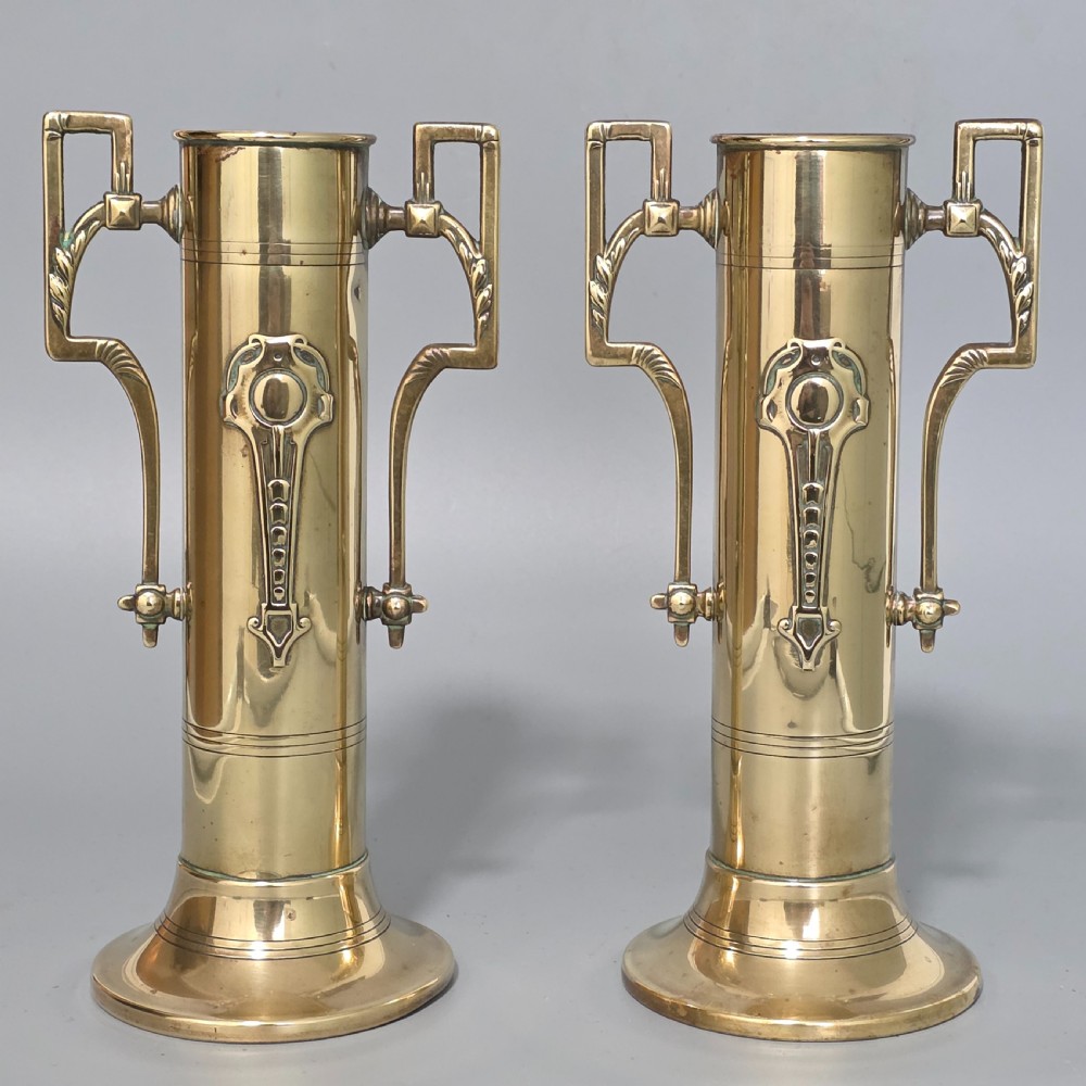 A pair of antique brass cylindrical vases featuring angular twin handles and raised stylised relief details on flared circular bases. View III