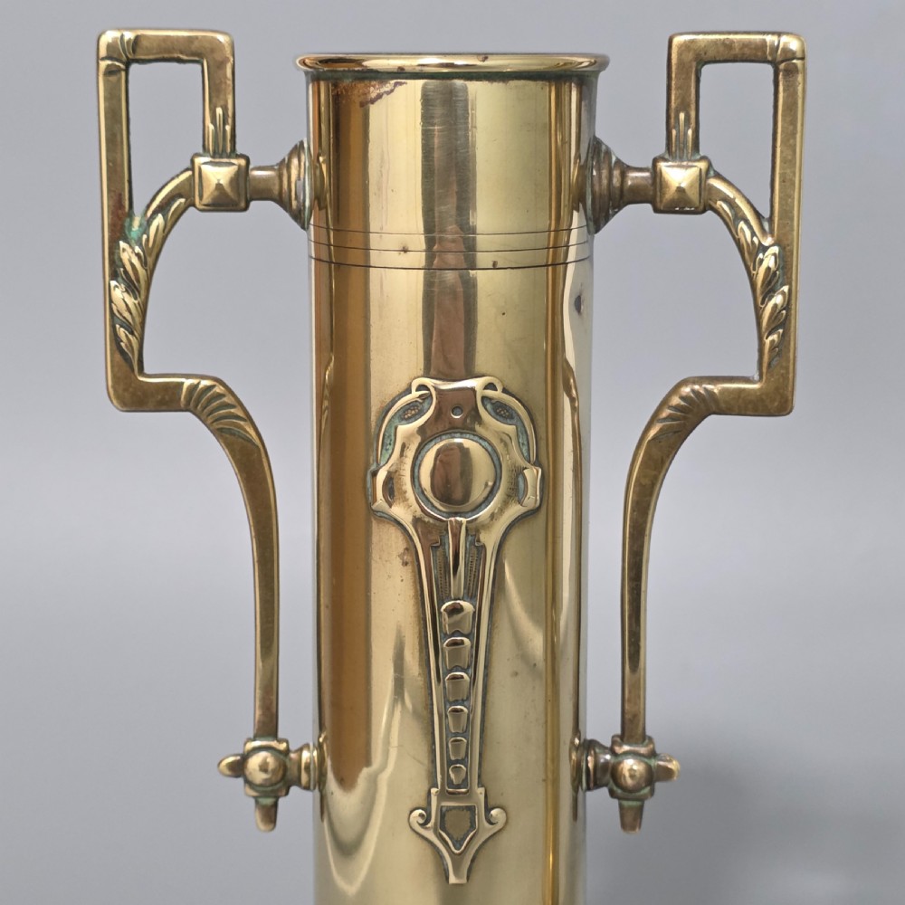 A pair of antique brass cylindrical vases featuring angular twin handles and raised stylised relief details on flared circular bases. View IV