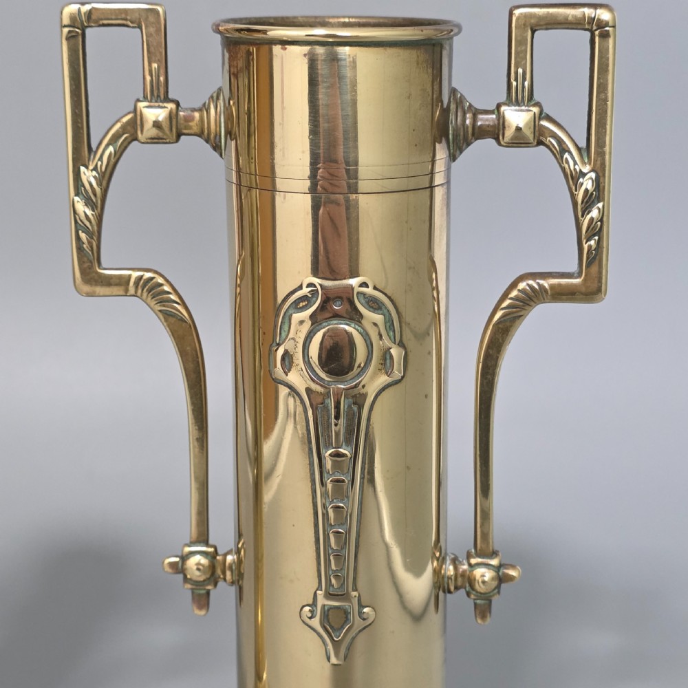 A pair of antique brass cylindrical vases featuring angular twin handles and raised stylised relief details on flared circular bases. View VI
