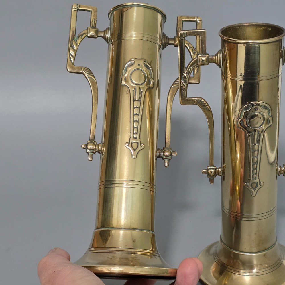 A pair of antique brass cylindrical vases featuring angular twin handles and raised stylised relief details on flared circular bases. View VII