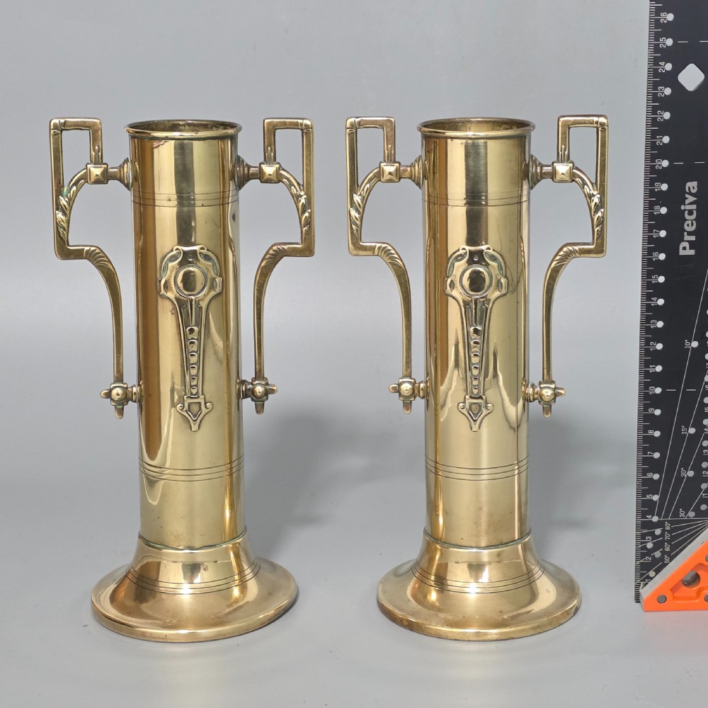 A pair of antique brass cylindrical vases featuring angular twin handles and raised stylised relief details on flared circular bases. View IX
