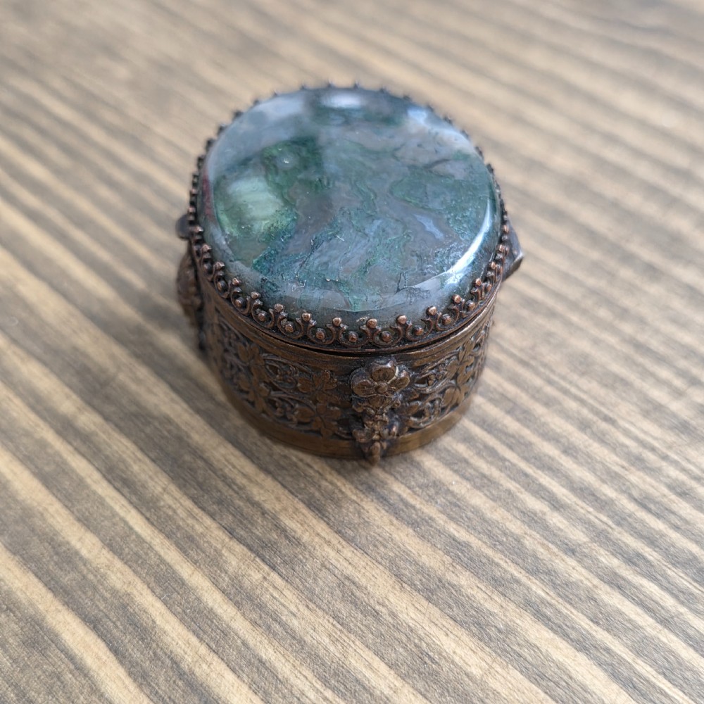 A small oval Victorian gilt metal pill box featuring a polished green moss agate cabochon lid and detailed relief scrollwork on the sides.