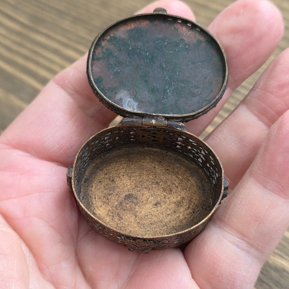 A small oval Victorian gilt metal pill box featuring a polished green moss agate cabochon lid and detailed relief scrollwork on the sides. View VI