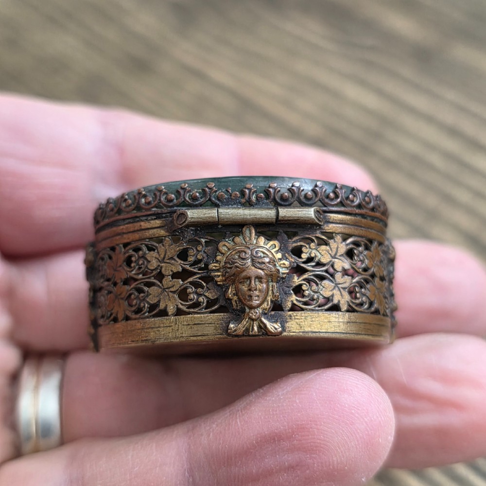 A small oval Victorian gilt metal pill box featuring a polished green moss agate cabochon lid and detailed relief scrollwork on the sides. View VII