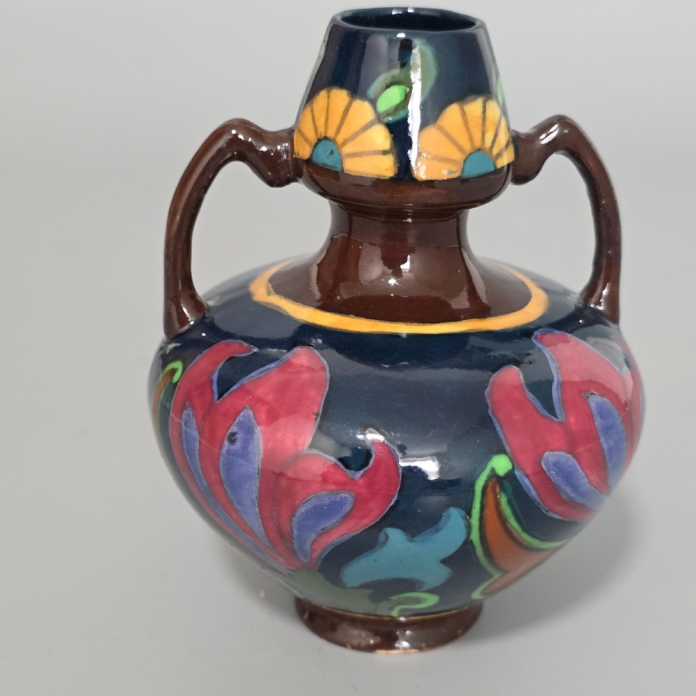 A glazed earthenware Art Nouveau vase with twin handles featuring a dark blue and brown ground decorated with stylised pink, purple, yellow, and green floral patterns.