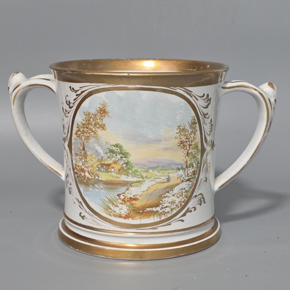 A nineteenth-century ceramic loving cup featuring two side handles, gilt trim, and a hand-painted landscape scene inside an oval cartouche.