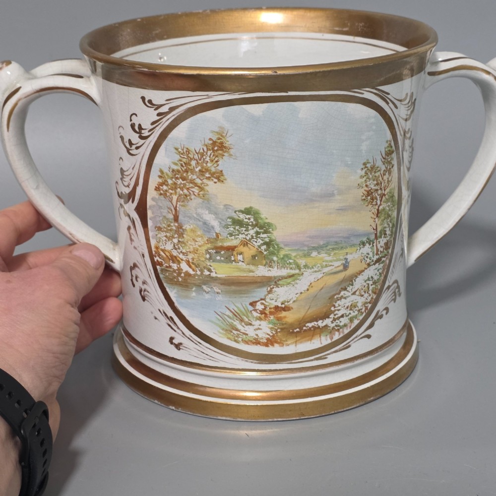A nineteenth-century ceramic loving cup featuring two side handles, gilt trim, and a hand-painted landscape scene inside an oval cartouche. View VI