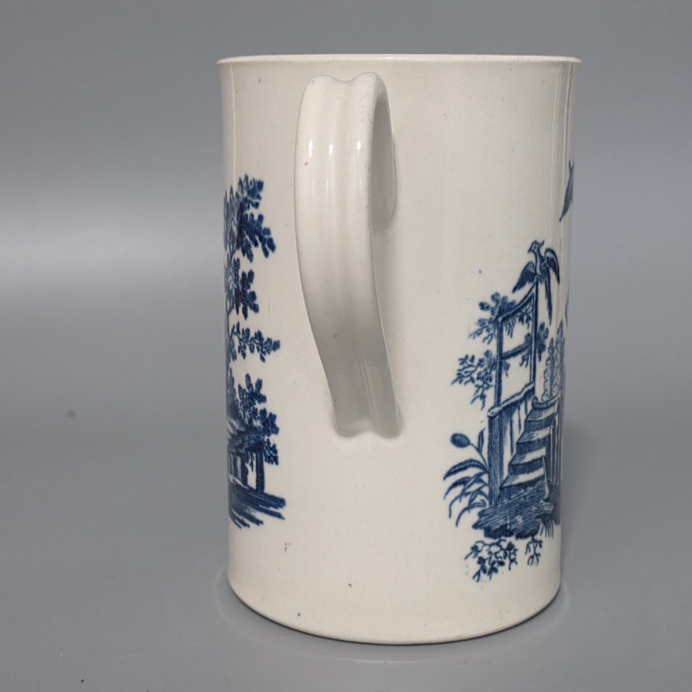 A tall cylindrical white porcelain mug featuring an 18th-century blue underglaze transfer-printed Chinoiserie design of figures and parasols with a single loop handle. View VI