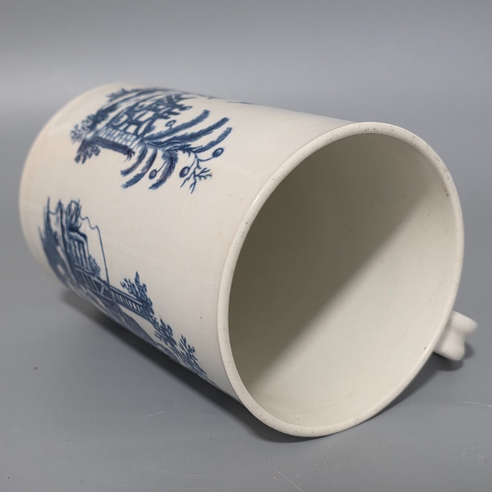 A tall cylindrical white porcelain mug featuring an 18th-century blue underglaze transfer-printed Chinoiserie design of figures and parasols with a single loop handle. View X