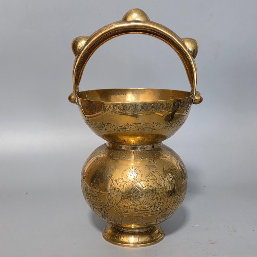 An antique Indian engraved brass kamandal vessel featuring a rounded base decorated with figures, a flared upper rim, and a high arched handle with raised knops.