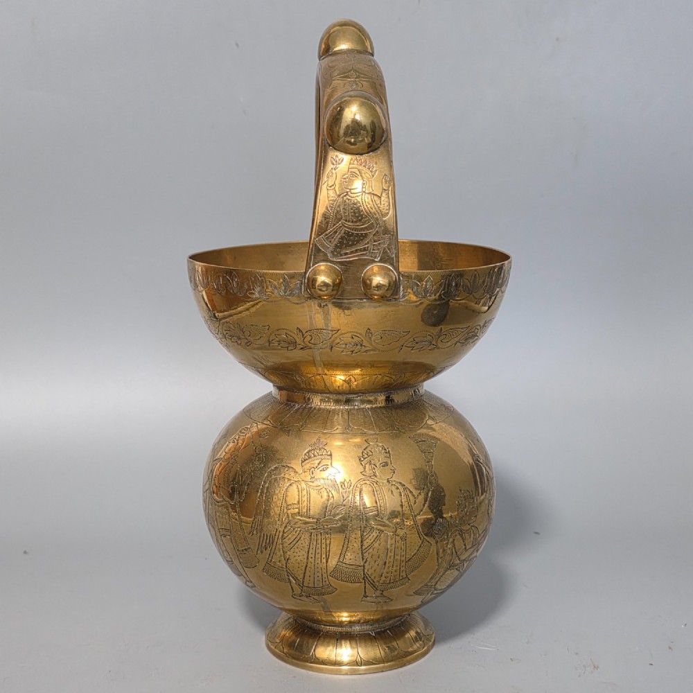 An antique Indian engraved brass kamandal vessel featuring a rounded base decorated with figures, a flared upper rim, and a high arched handle with raised knops. View II