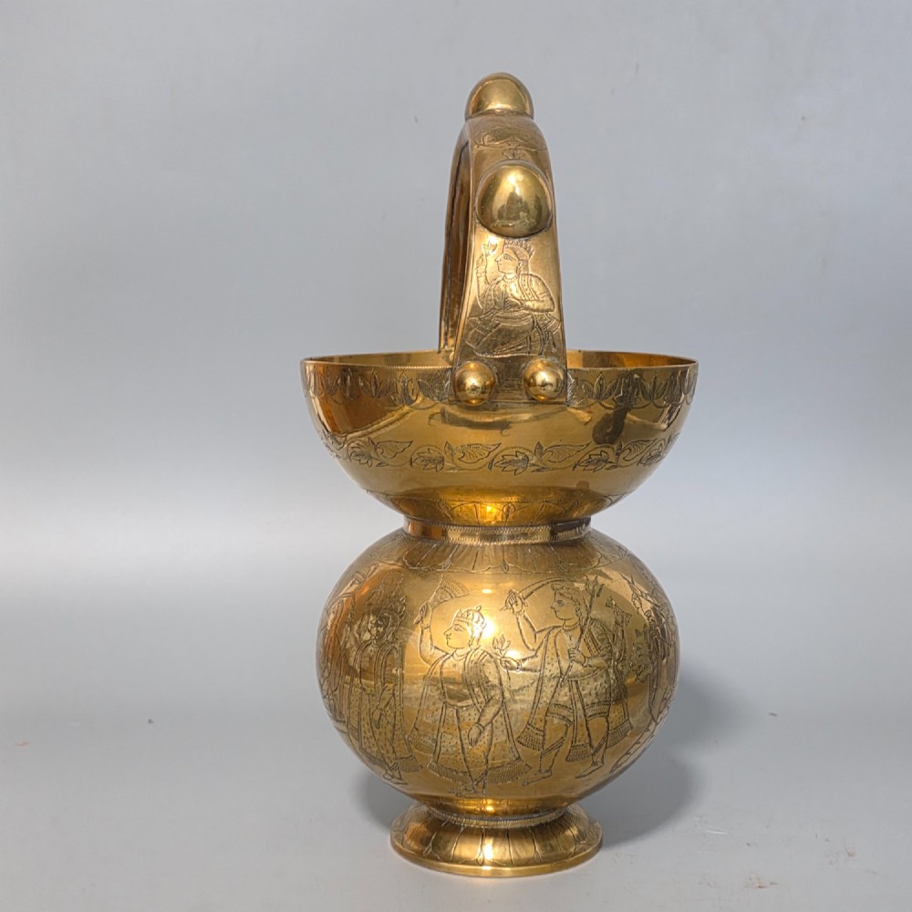 An antique Indian engraved brass kamandal vessel featuring a rounded base decorated with figures, a flared upper rim, and a high arched handle with raised knops. View III