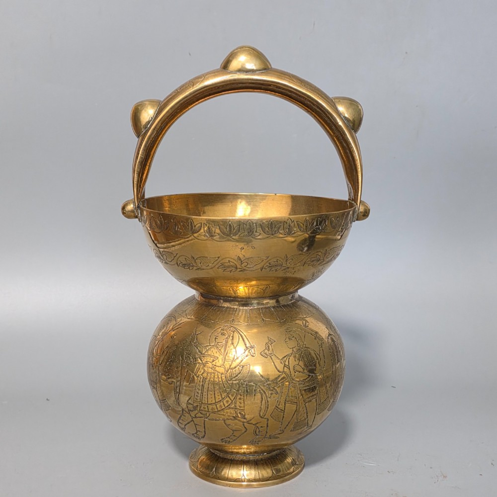 An antique Indian engraved brass kamandal vessel featuring a rounded base decorated with figures, a flared upper rim, and a high arched handle with raised knops. View X