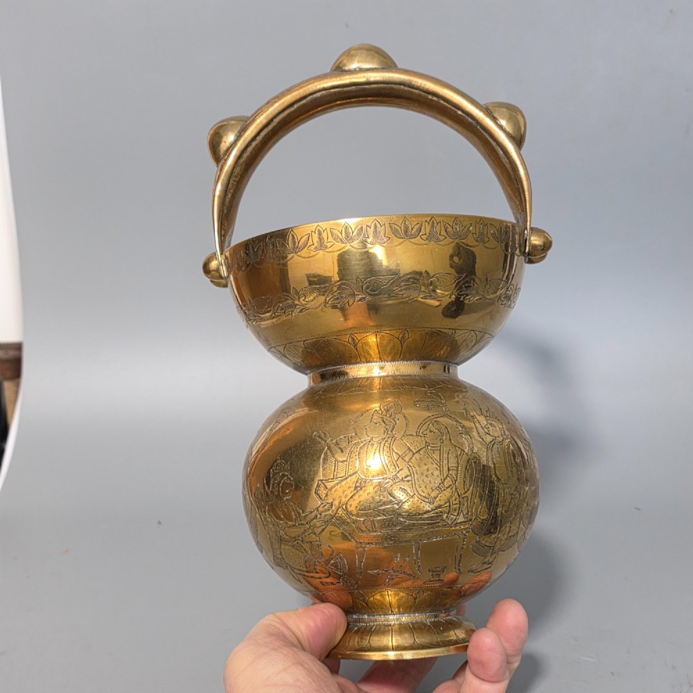 An antique Indian engraved brass kamandal vessel featuring a rounded base decorated with figures, a flared upper rim, and a high arched handle with raised knops. View XIII
