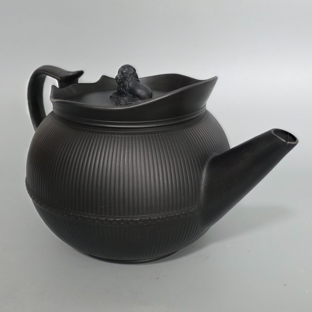 An 18th-century matte black basalt teapot featuring vertical engine-turned fluting, a curved spout, and a lid crowned with a sculpted lion finial.