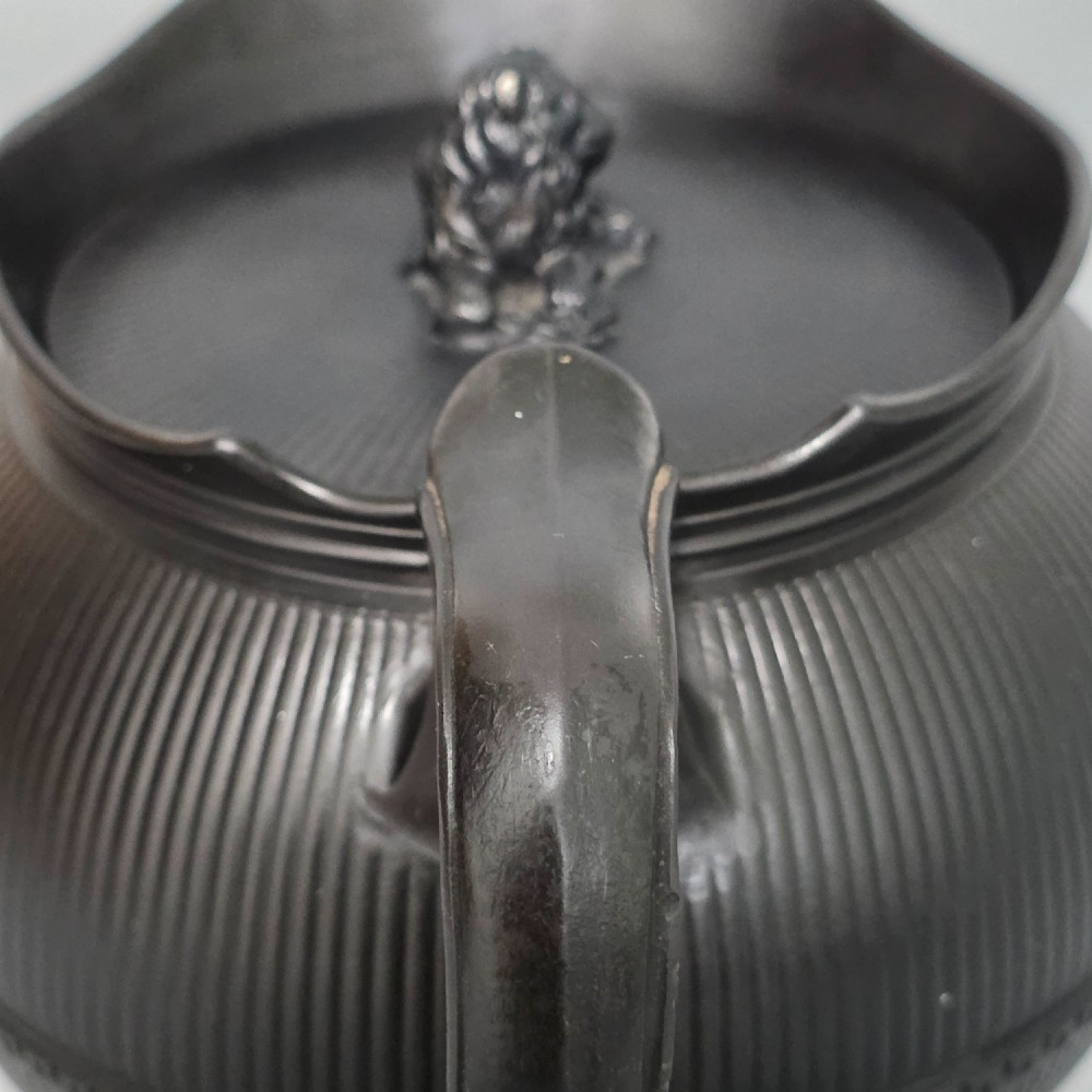 An 18th-century matte black basalt teapot featuring vertical engine-turned fluting, a curved spout, and a lid crowned with a sculpted lion finial. View VII