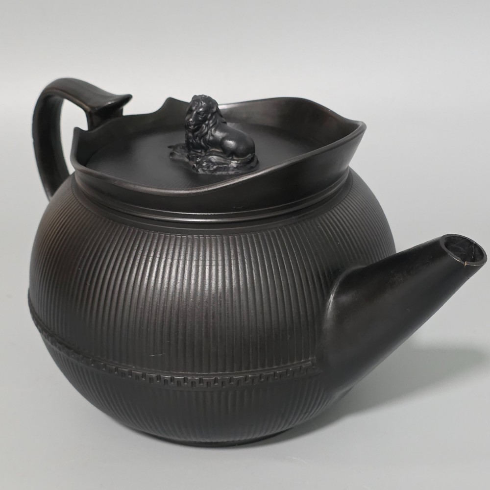 An 18th-century matte black basalt teapot featuring vertical engine-turned fluting, a curved spout, and a lid crowned with a sculpted lion finial. View X