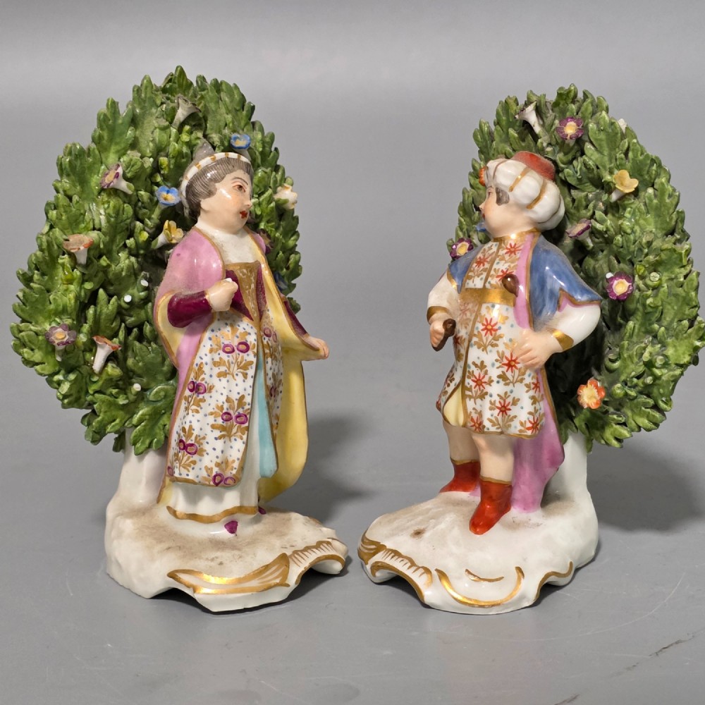 A pair of 19th-century hand-painted porcelain figurines featuring male and female characters in detailed costume before dense floral bocage backdrops on gilded scroll bases.