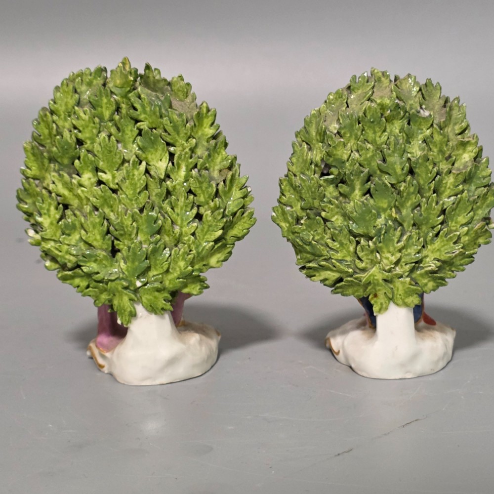A pair of 19th-century hand-painted porcelain figurines featuring male and female characters in detailed costume before dense floral bocage backdrops on gilded scroll bases. View II