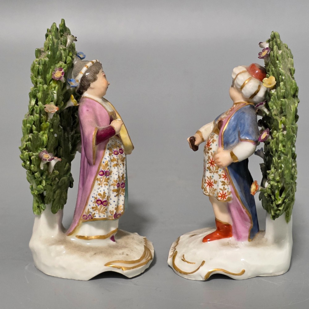 A pair of 19th-century hand-painted porcelain figurines featuring male and female characters in detailed costume before dense floral bocage backdrops on gilded scroll bases. View III