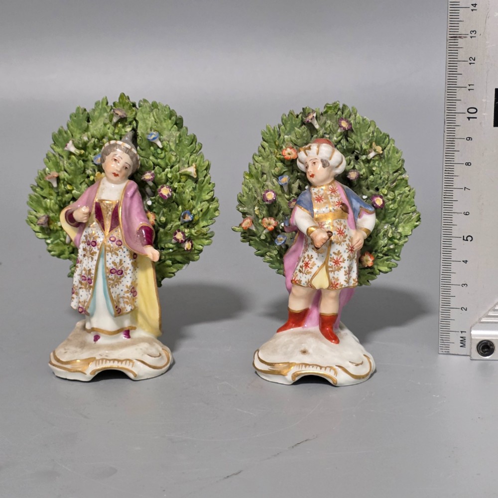 A pair of 19th-century hand-painted porcelain figurines featuring male and female characters in detailed costume before dense floral bocage backdrops on gilded scroll bases. View IV
