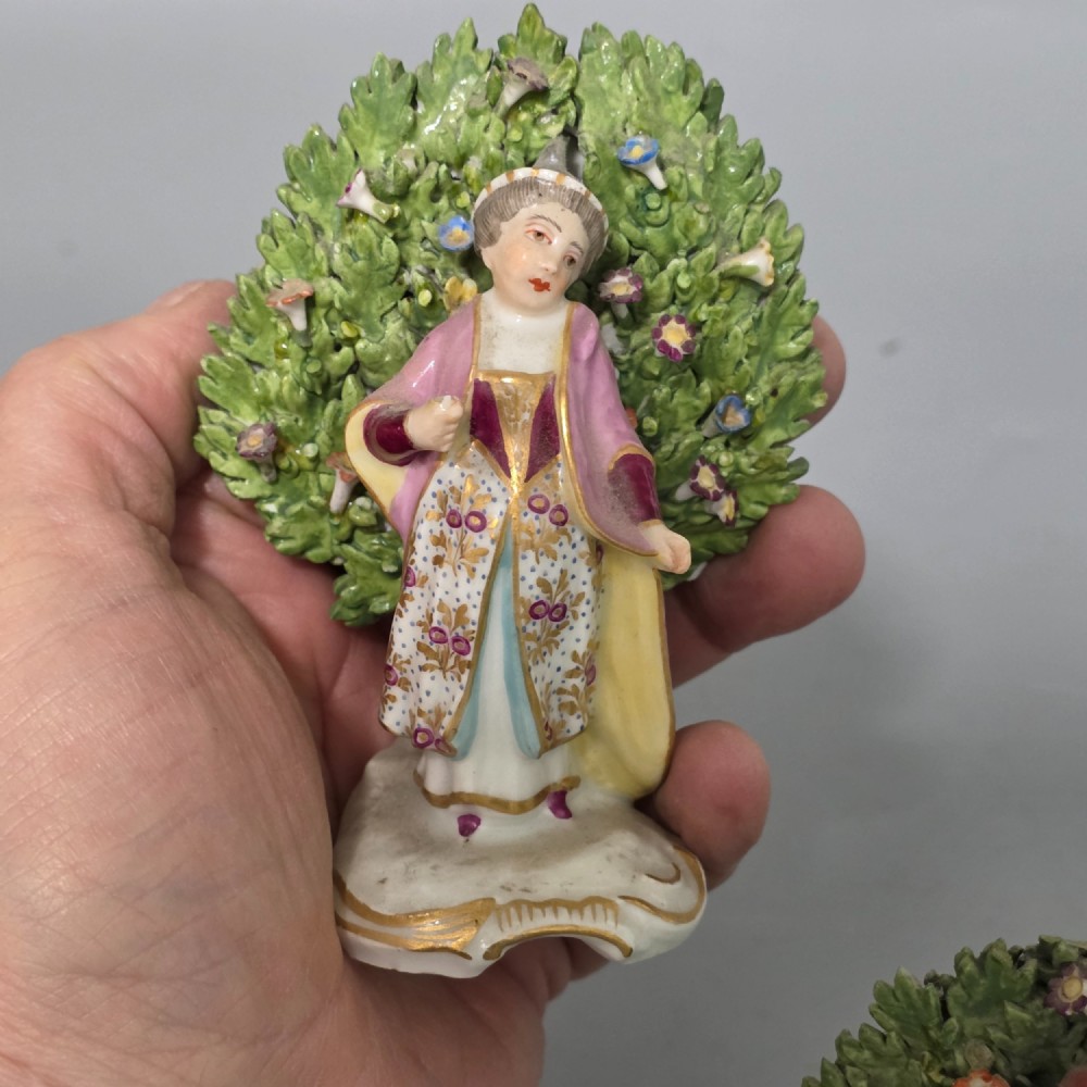 A pair of 19th-century hand-painted porcelain figurines featuring male and female characters in detailed costume before dense floral bocage backdrops on gilded scroll bases. View V