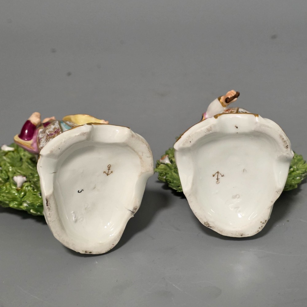 A pair of 19th-century hand-painted porcelain figurines featuring male and female characters in detailed costume before dense floral bocage backdrops on gilded scroll bases. View XI