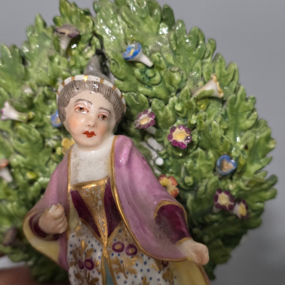 A pair of 19th-century hand-painted porcelain figurines featuring male and female characters in detailed costume before dense floral bocage backdrops on gilded scroll bases. View XII