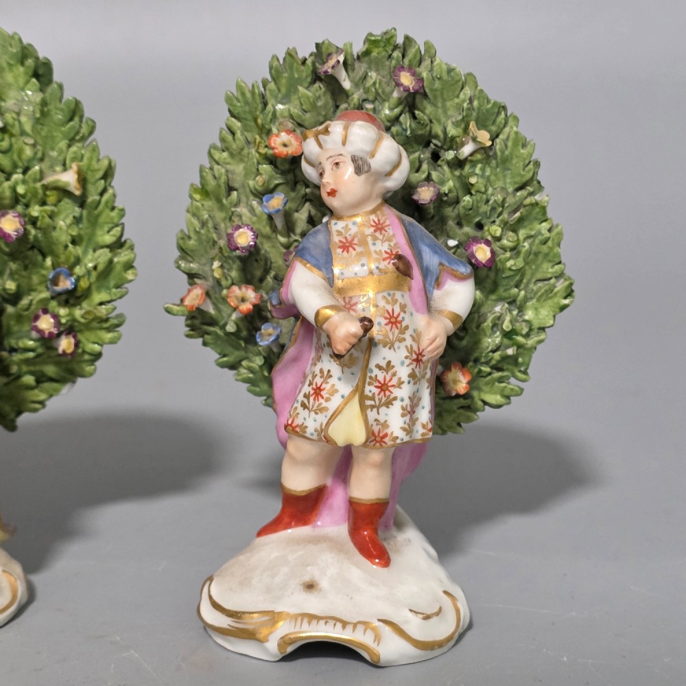 A pair of 19th-century hand-painted porcelain figurines featuring male and female characters in detailed costume before dense floral bocage backdrops on gilded scroll bases. View XIII