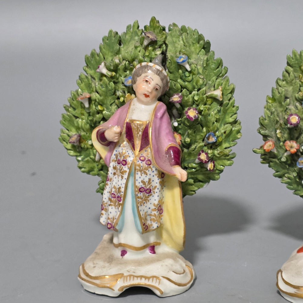 A pair of 19th-century hand-painted porcelain figurines featuring male and female characters in detailed costume before dense floral bocage backdrops on gilded scroll bases. View XIV