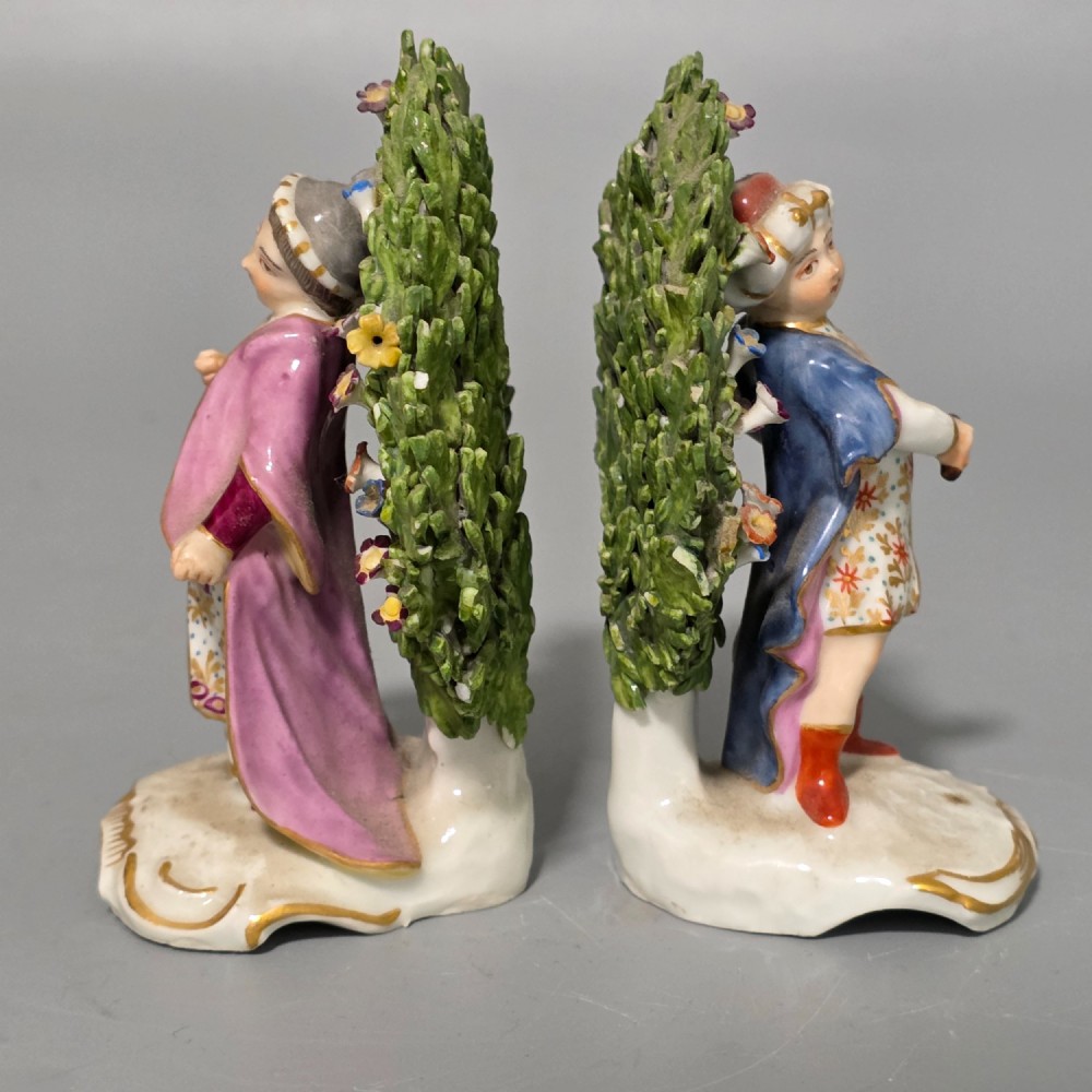 A pair of 19th-century hand-painted porcelain figurines featuring male and female characters in detailed costume before dense floral bocage backdrops on gilded scroll bases. View XV