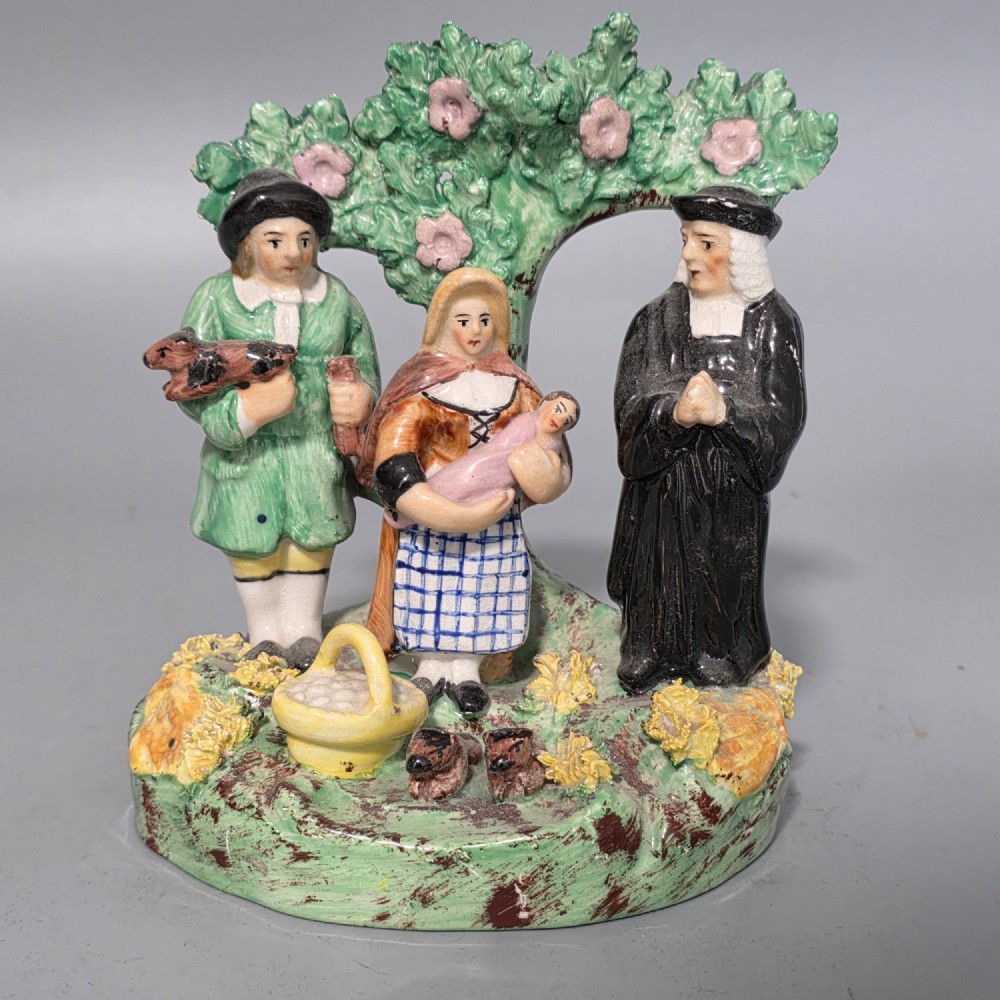 An early 19th-century Staffordshire ceramic figure group depicting a farmer, his wife holding a baby, and a parson standing before a flowering bocage tree.