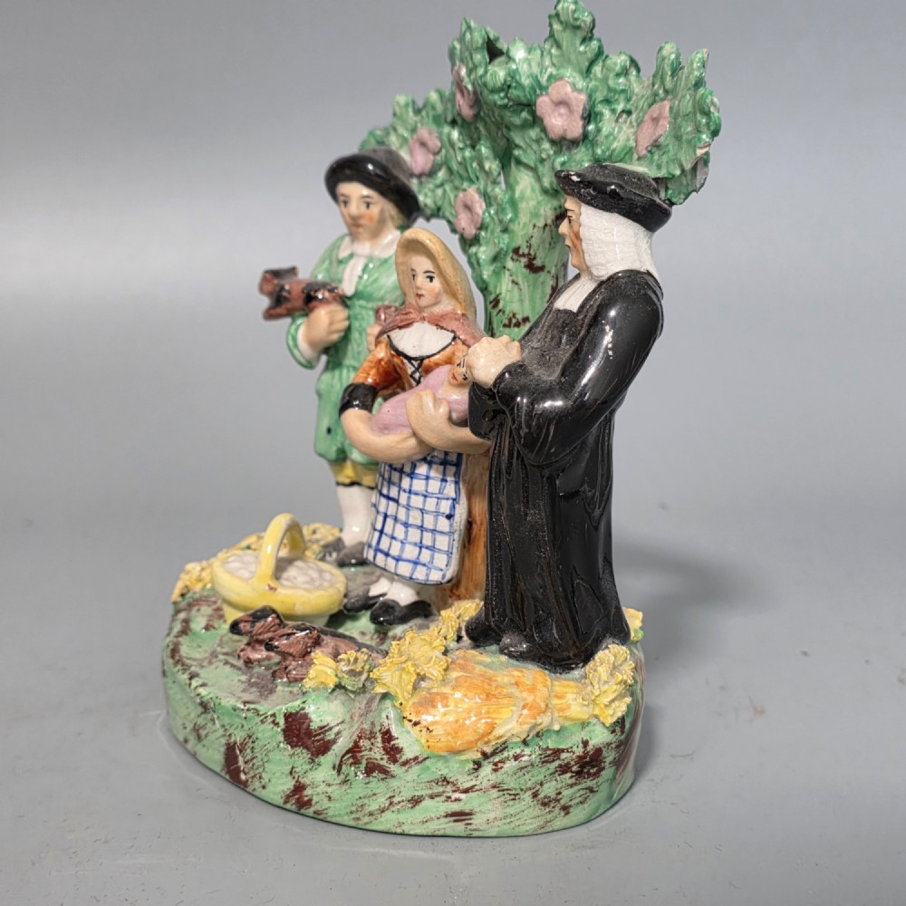 An early 19th-century Staffordshire ceramic figure group depicting a farmer, his wife holding a baby, and a parson standing before a flowering bocage tree. View IX