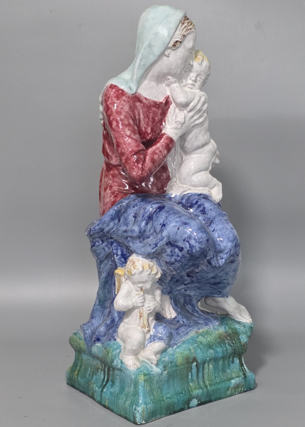 A polychrome glazed ceramic sculpture depicting an enthroned Madonna holding the Christ Child with a small musical putto playing at her feet.