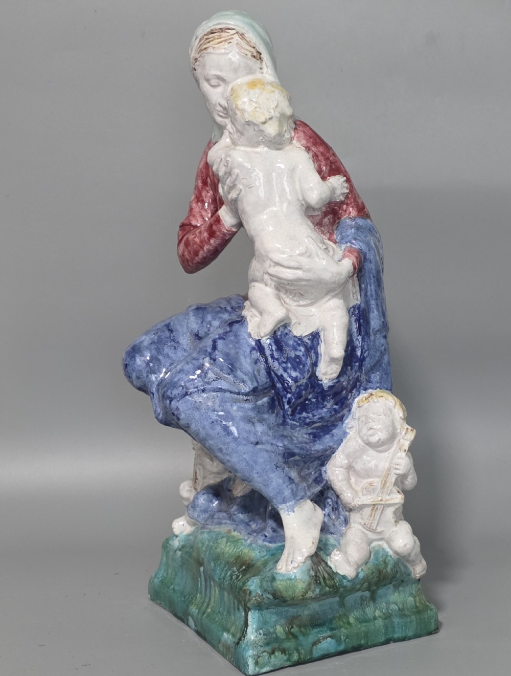 A polychrome glazed ceramic sculpture depicting an enthroned Madonna holding the Christ Child with a small musical putto playing at her feet. View III