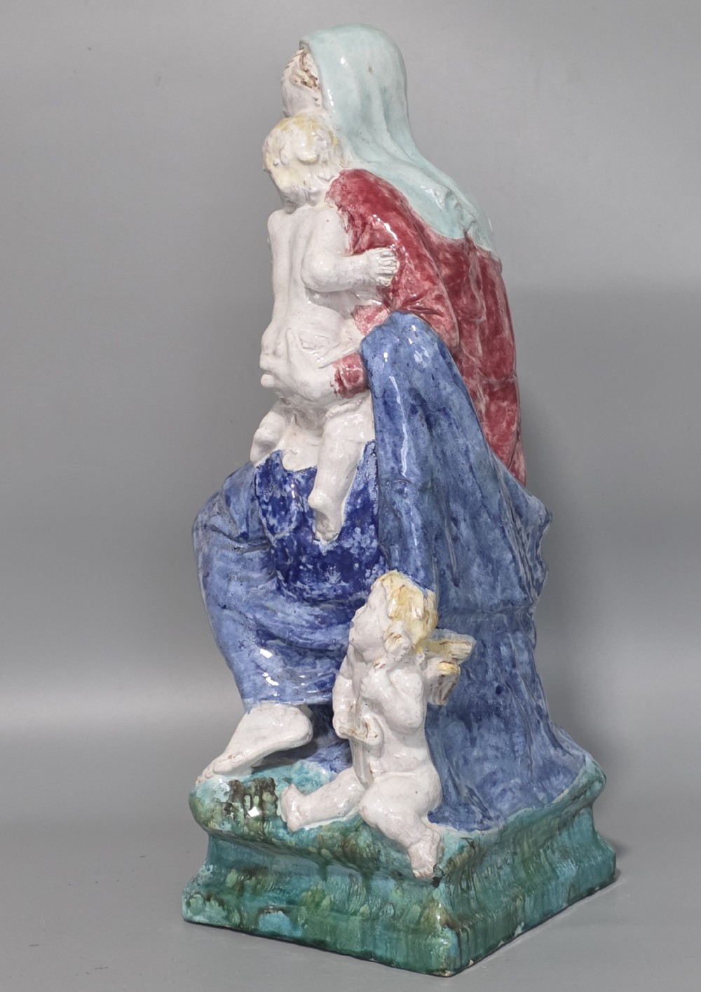 A polychrome glazed ceramic sculpture depicting an enthroned Madonna holding the Christ Child with a small musical putto playing at her feet. View IV