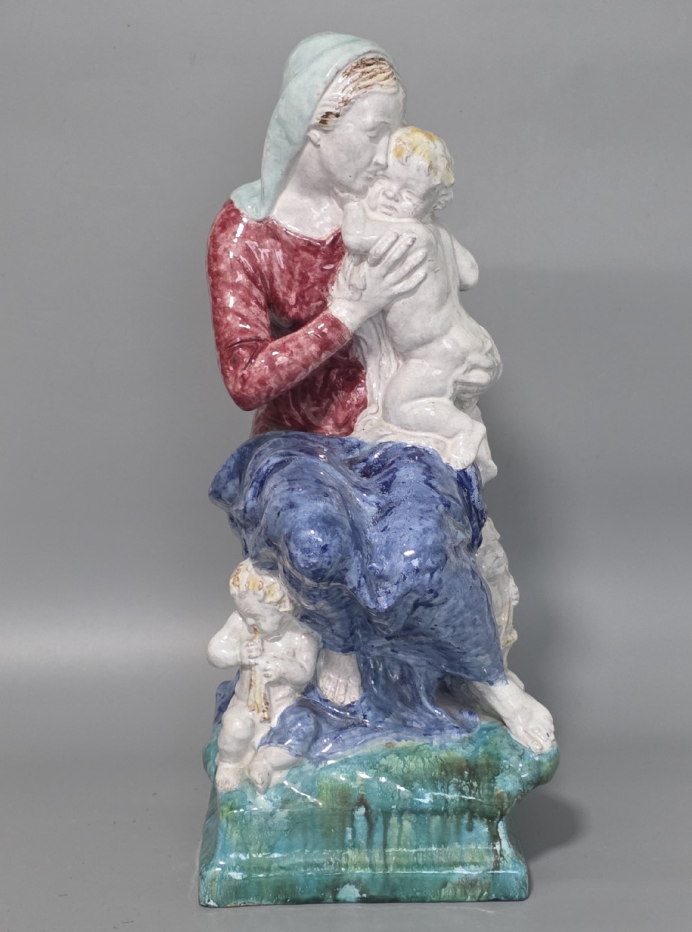 A polychrome glazed ceramic sculpture depicting an enthroned Madonna holding the Christ Child with a small musical putto playing at her feet. View VIII