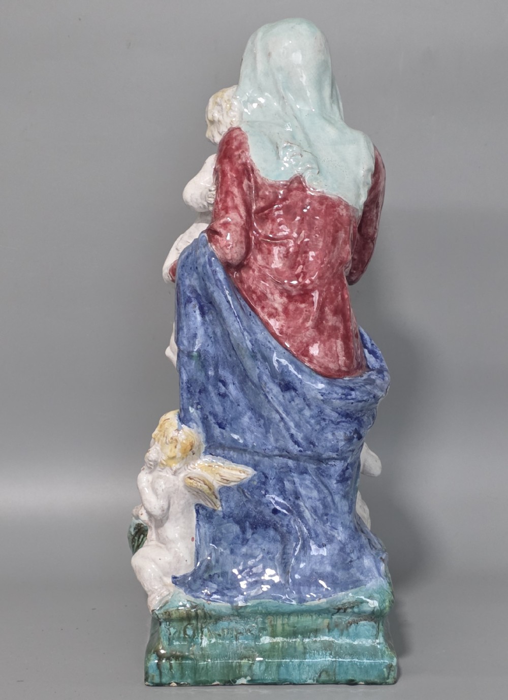 A polychrome glazed ceramic sculpture depicting an enthroned Madonna holding the Christ Child with a small musical putto playing at her feet. View IX
