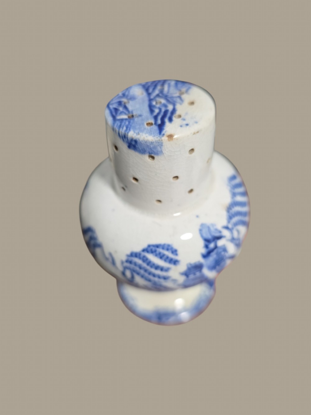 A small antique blue and white pearlware pepper pot with pierced holes on top and a transferware landscape pattern.