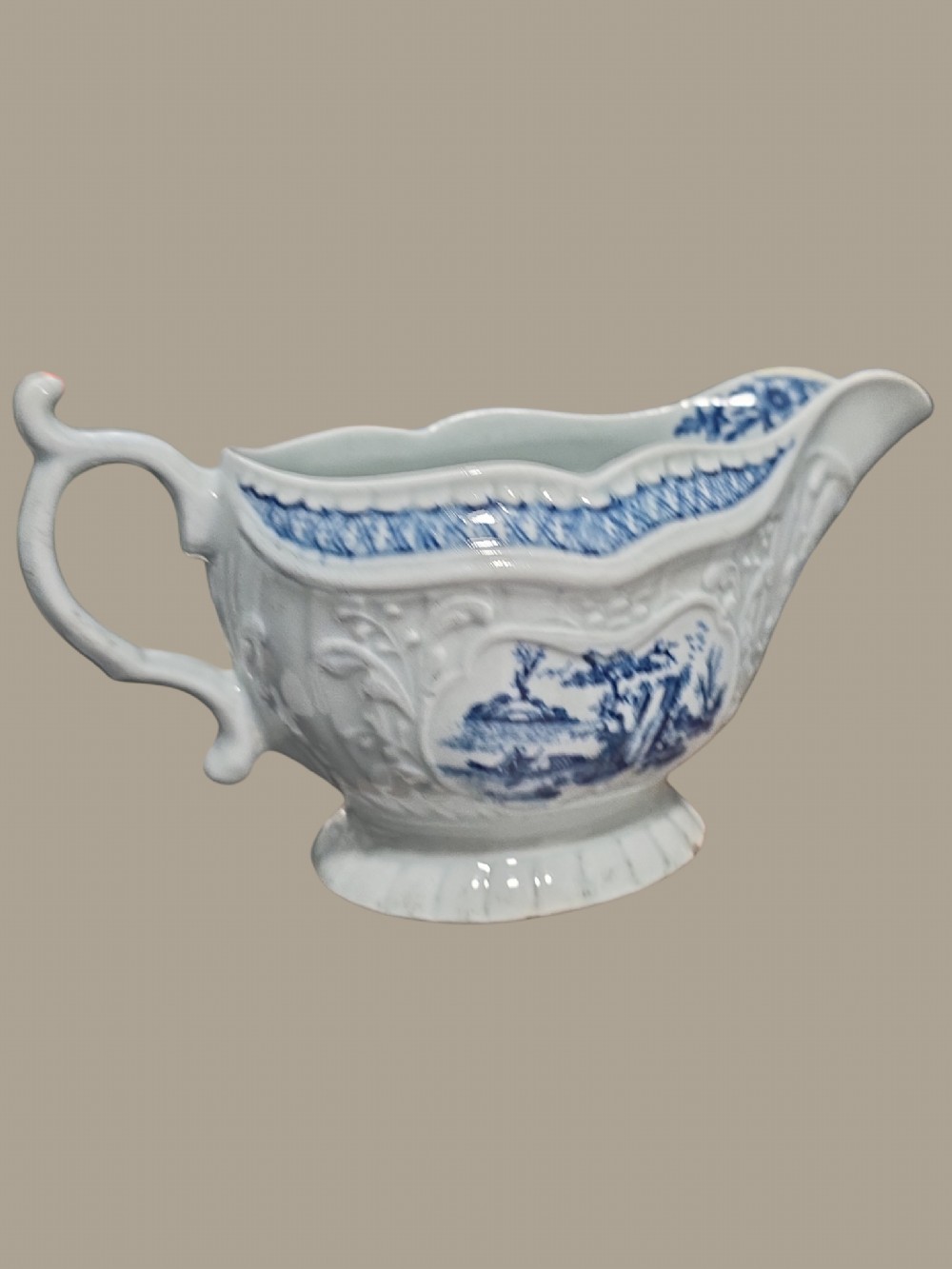 An eighteenth-century blue and white soft-paste porcelain sauce boat featuring moulded scroll decoration, a shaped rim, a scroll handle, and a printed landscape scene.