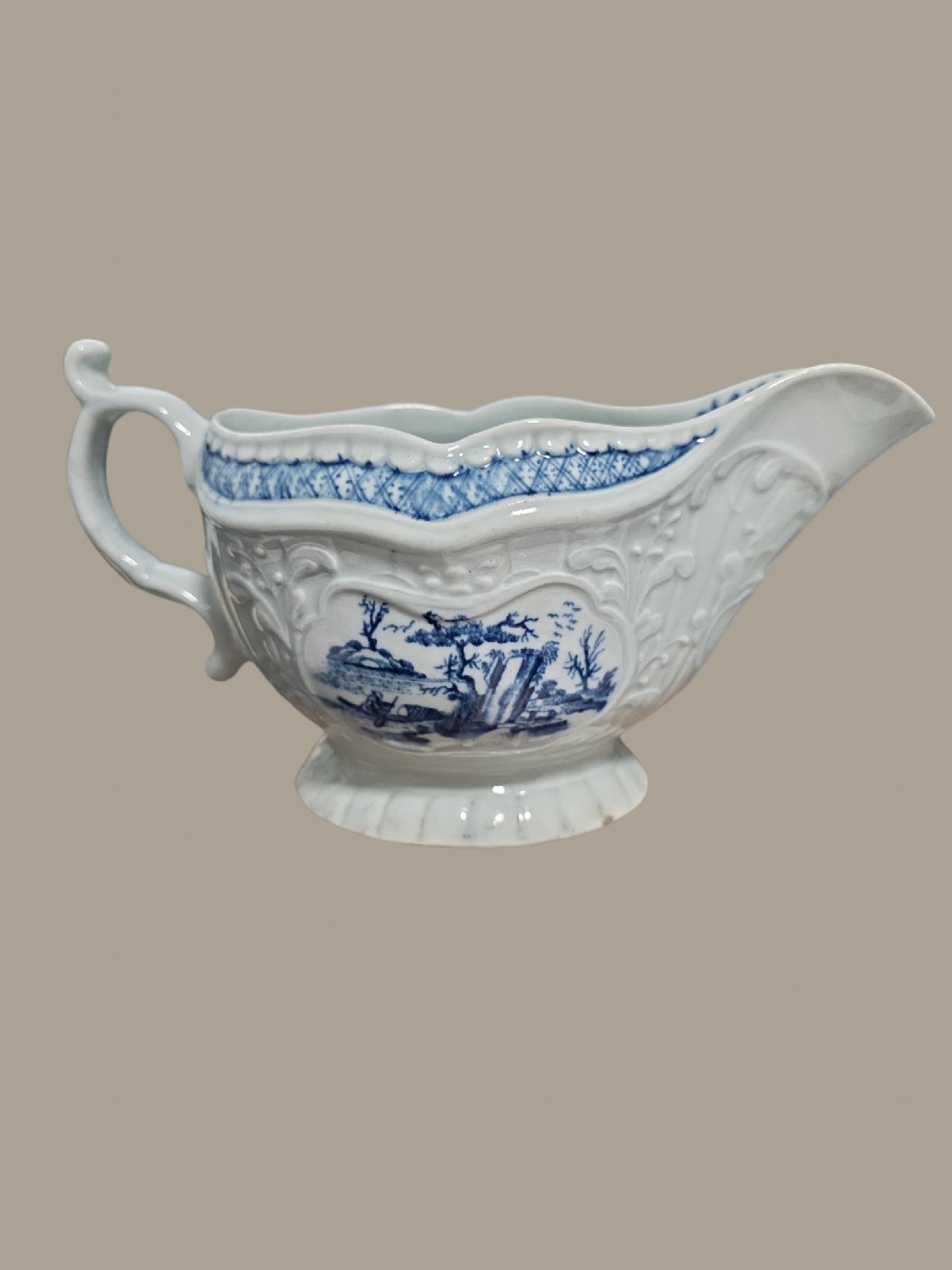 An eighteenth-century blue and white soft-paste porcelain sauce boat featuring moulded scroll decoration, a shaped rim, a scroll handle, and a printed landscape scene. View VI