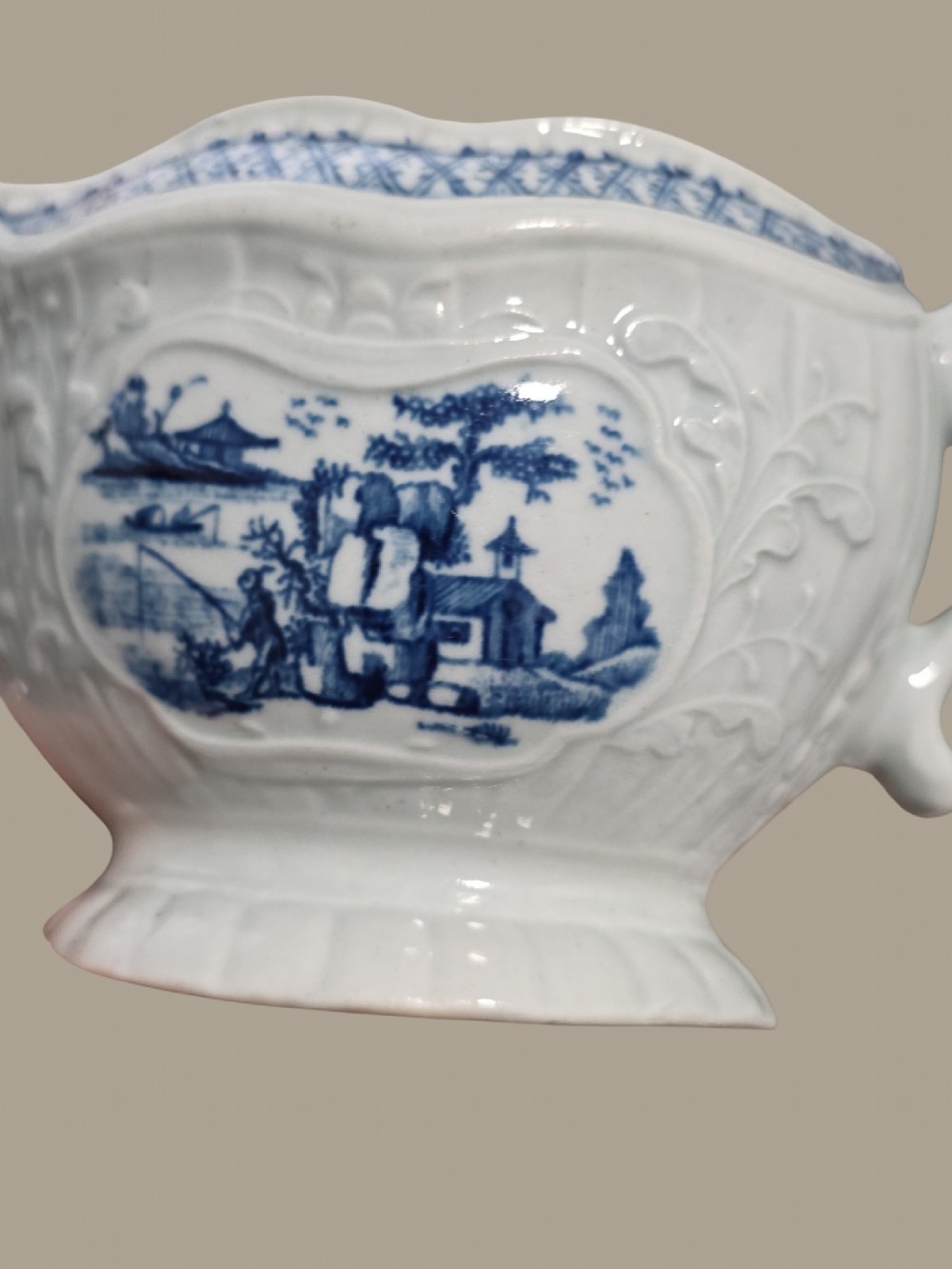 An eighteenth-century blue and white soft-paste porcelain sauce boat featuring moulded scroll decoration, a shaped rim, a scroll handle, and a printed landscape scene. View X