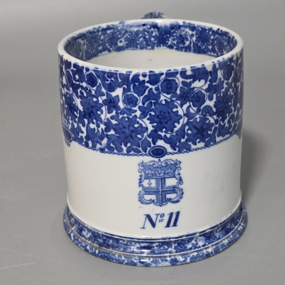 A cylindrical blue and white transferware ceramic tankard featuring a floral upper border and a coat of arms crest with the text No 11.