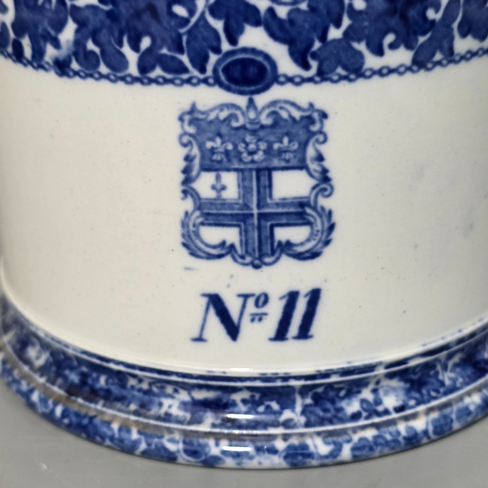 A cylindrical blue and white transferware ceramic tankard featuring a floral upper border and a coat of arms crest with the text No 11. View III