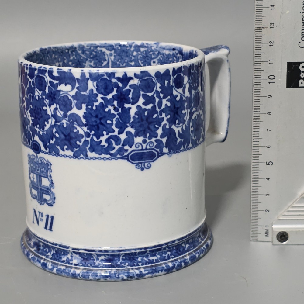 A cylindrical blue and white transferware ceramic tankard featuring a floral upper border and a coat of arms crest with the text No 11. View VI