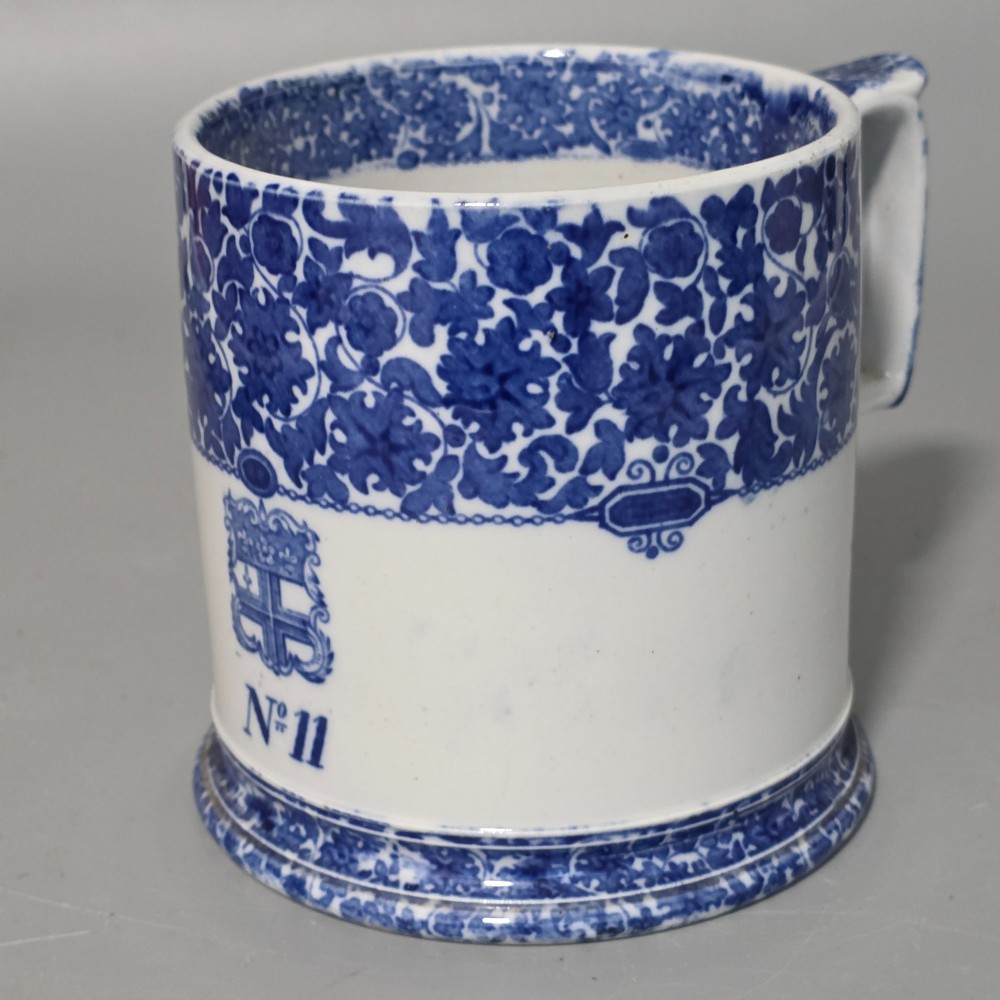 A cylindrical blue and white transferware ceramic tankard featuring a floral upper border and a coat of arms crest with the text No 11. View VII